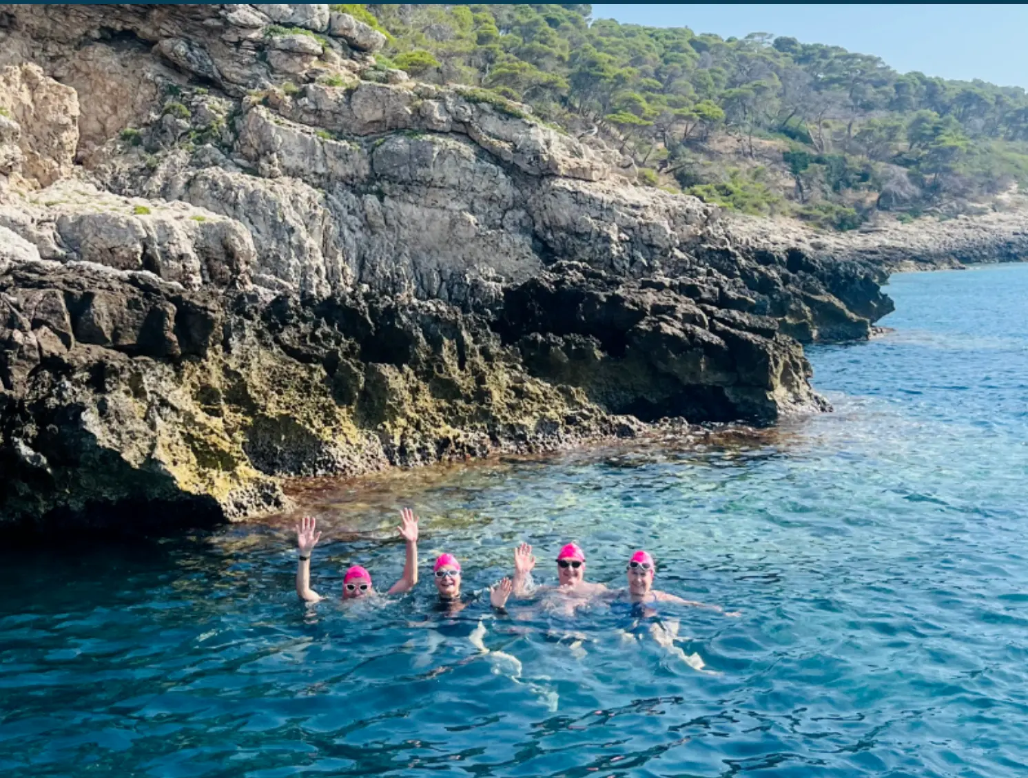 Group of swimmers waving in a clear turquoise cove beside limestone cliffs